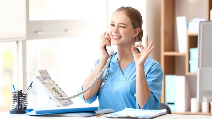 Person in blue scrubs sitting at a desk, holding a phone to their ear and making an "OK" gesture. Office supplies and shelves with books are visible in the background.